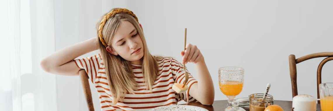 Teen girl in striped shirt at breakfast table with fruits, bread, and juice. Casual morning setting.